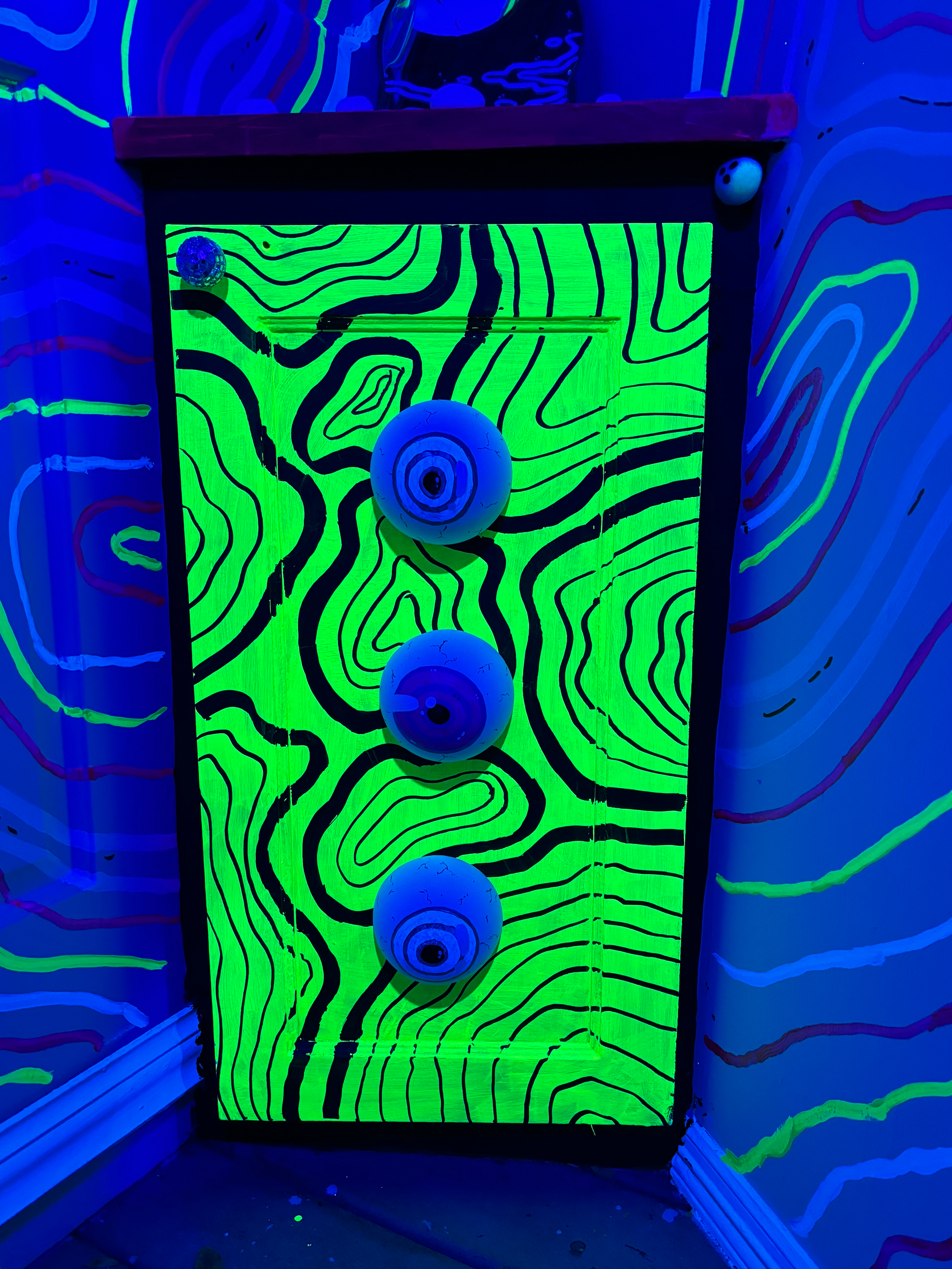 An abstract installation piece features three blue spheres against a neon green and black topographic pattern, illuminated by blacklight.