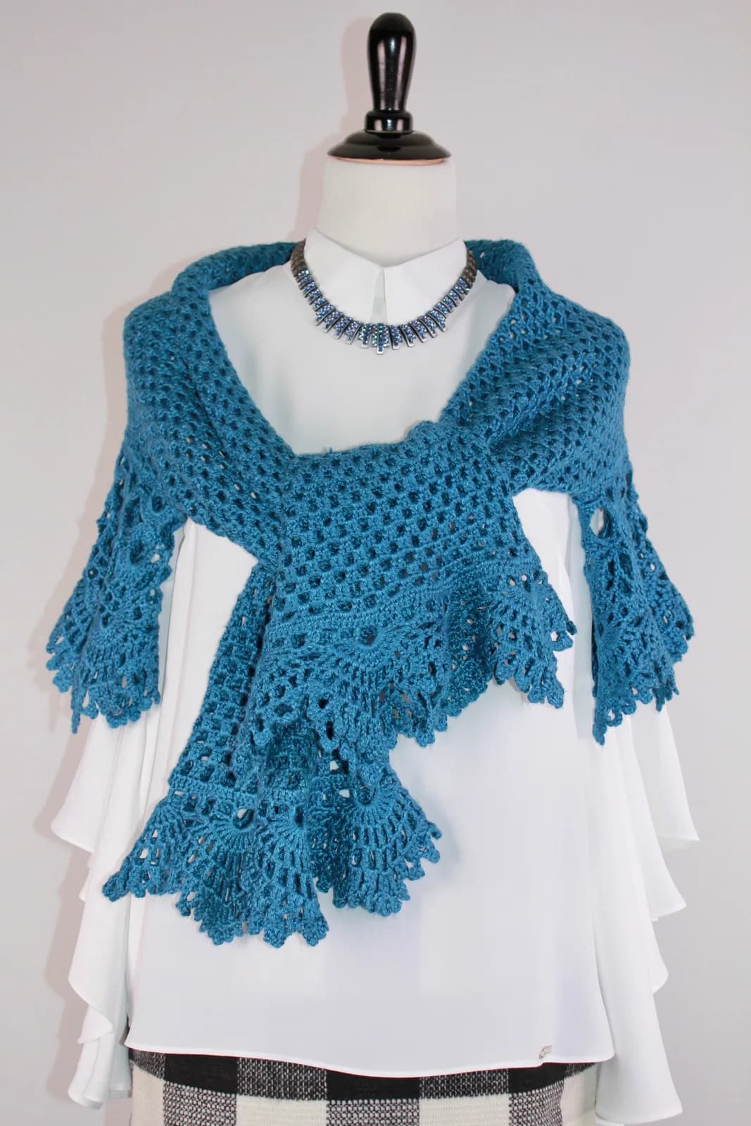 A blue crocheted shawl with intricate patterns is displayed over a white blouse, accessorized with a beaded necklace.