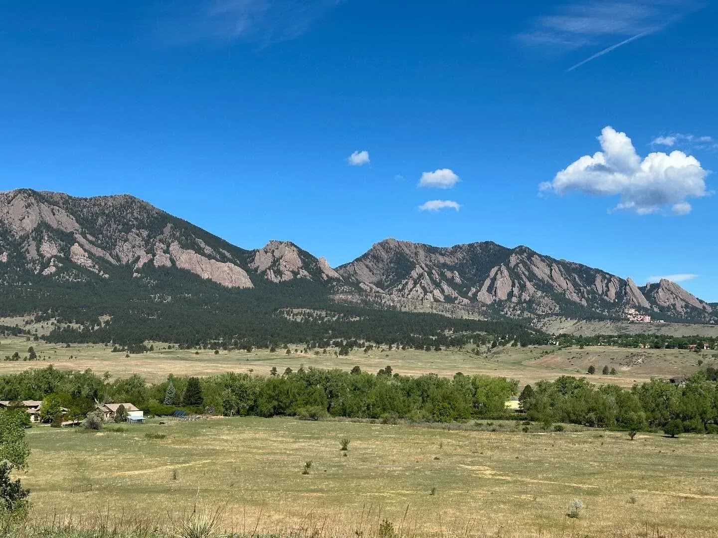 A landscape photograph captures the iconic Flatirons mountains under a bright blue sky with scattered clouds.