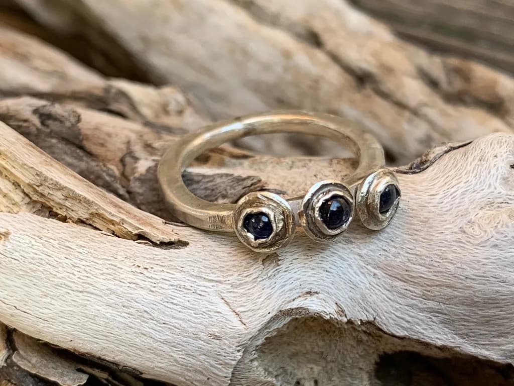 A metal ring with three dark blue stones is displayed on a piece of driftwood.
