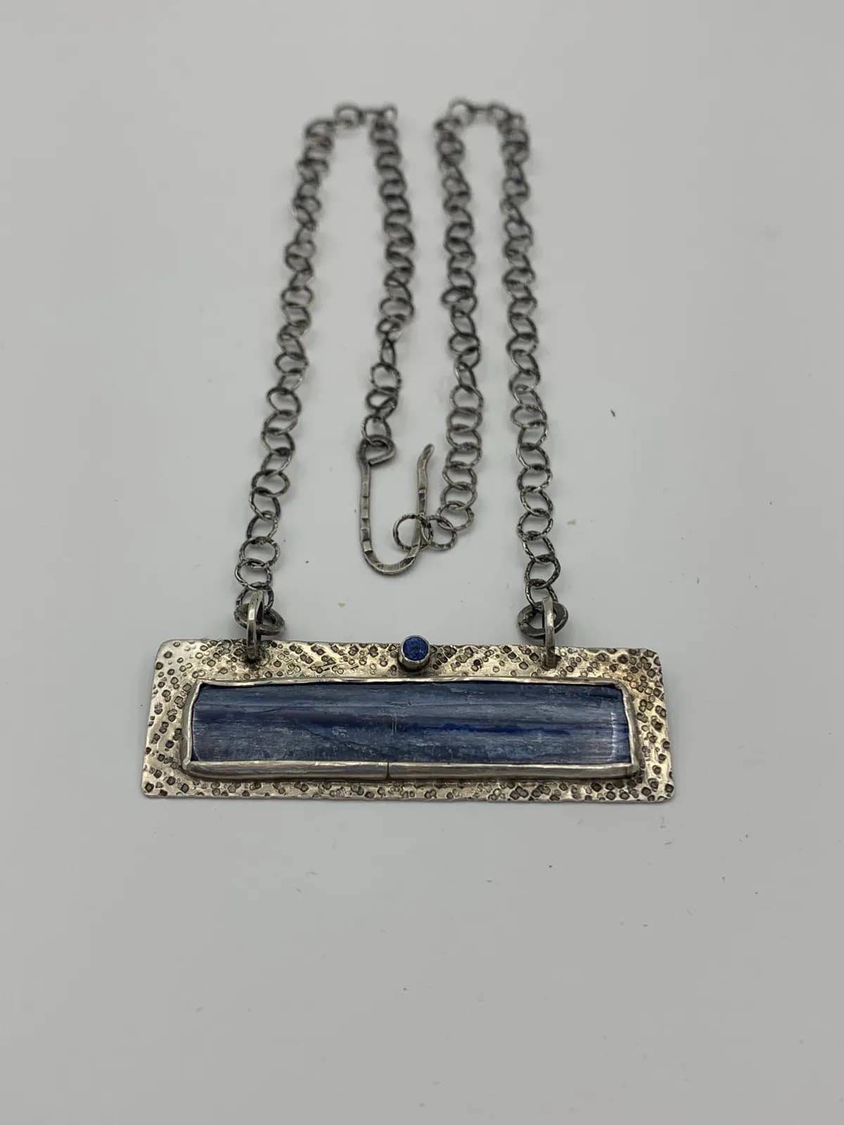 A rectangular silver pendant necklace features a blue stone and a textured metal frame.