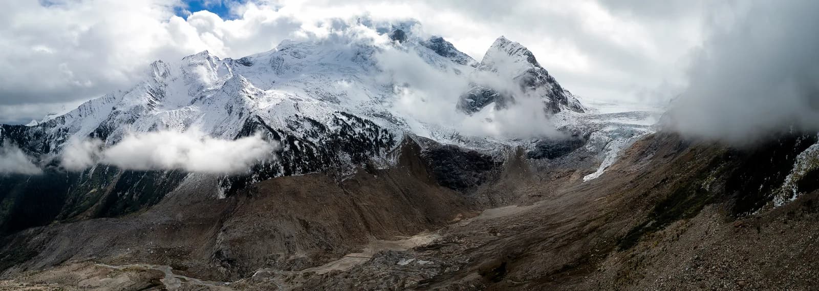 A panoramic photograph captures a majestic snow-capped mountain range shrouded in dramatic clouds, showcasing rocky terrain.