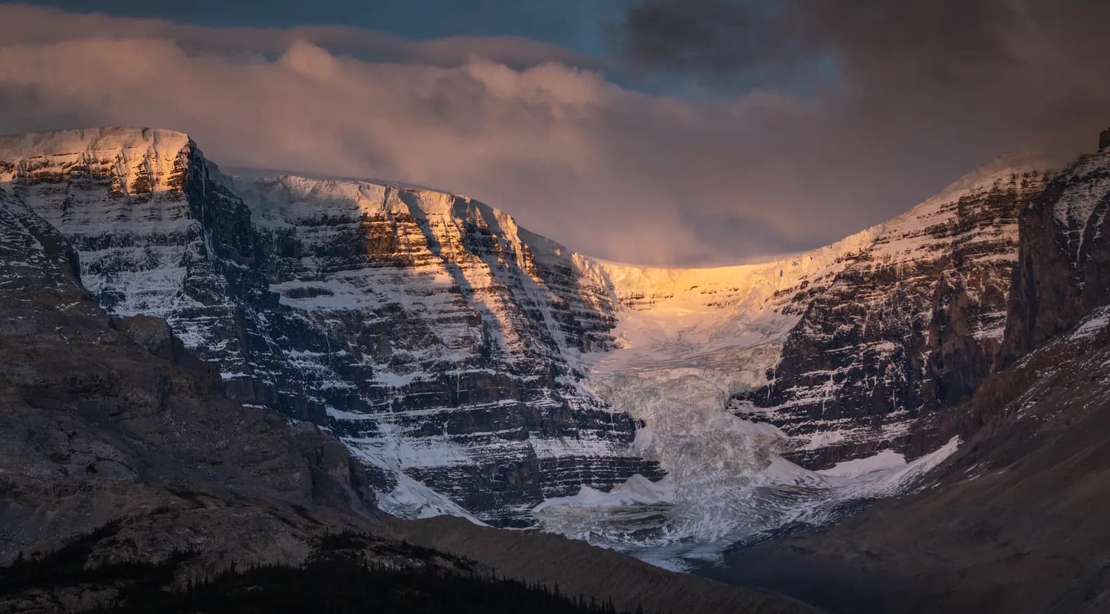 A dramatic sunset illuminates a snow-covered mountain range with a prominent glacier.