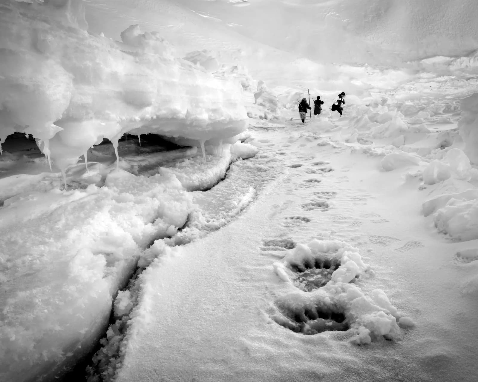 A black and white photograph captures a winter landscape with footprints in the snow and ice, leading towards distant figures.