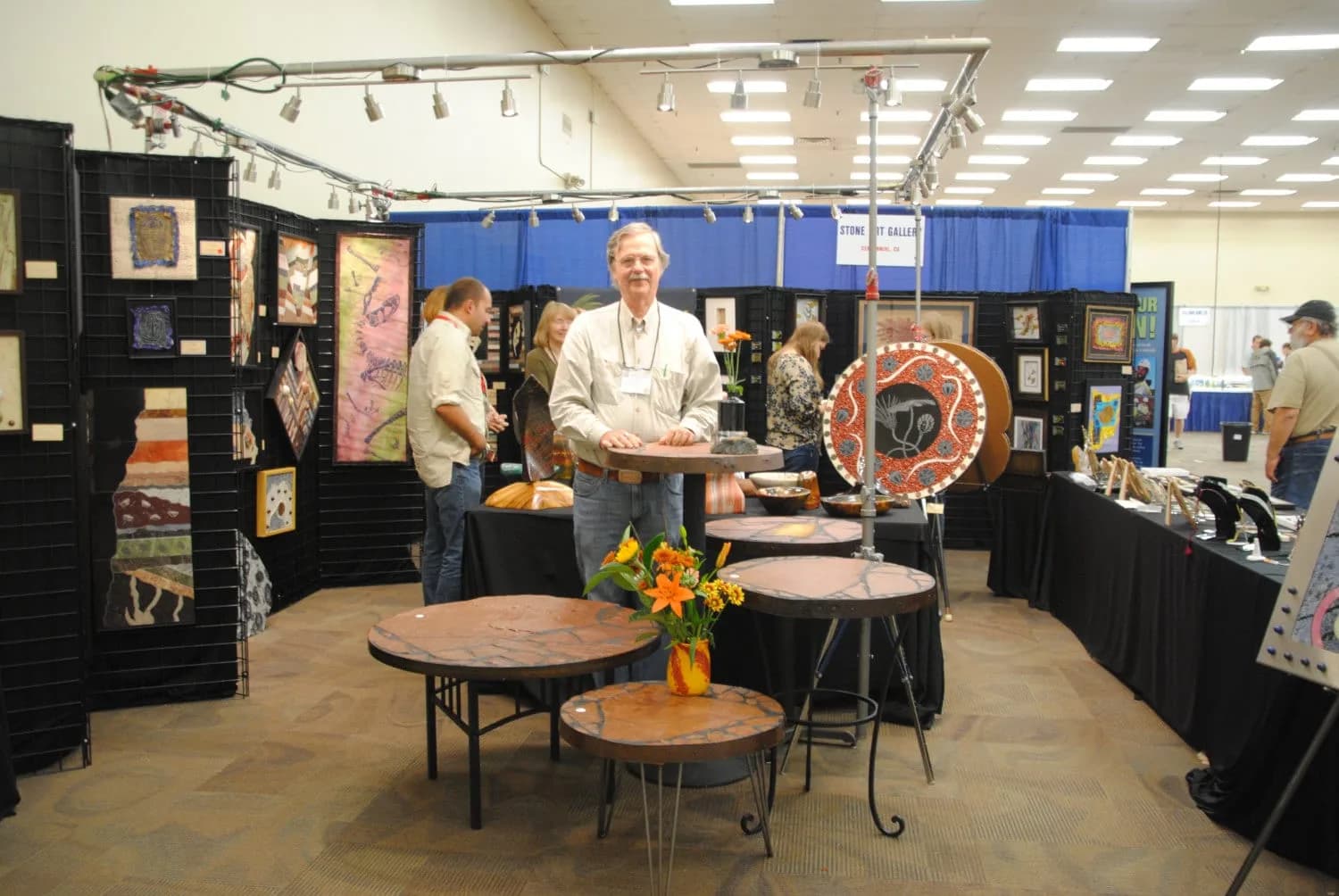 An artist stands in his booth at an art fair, surrounded by various artworks and display tables.