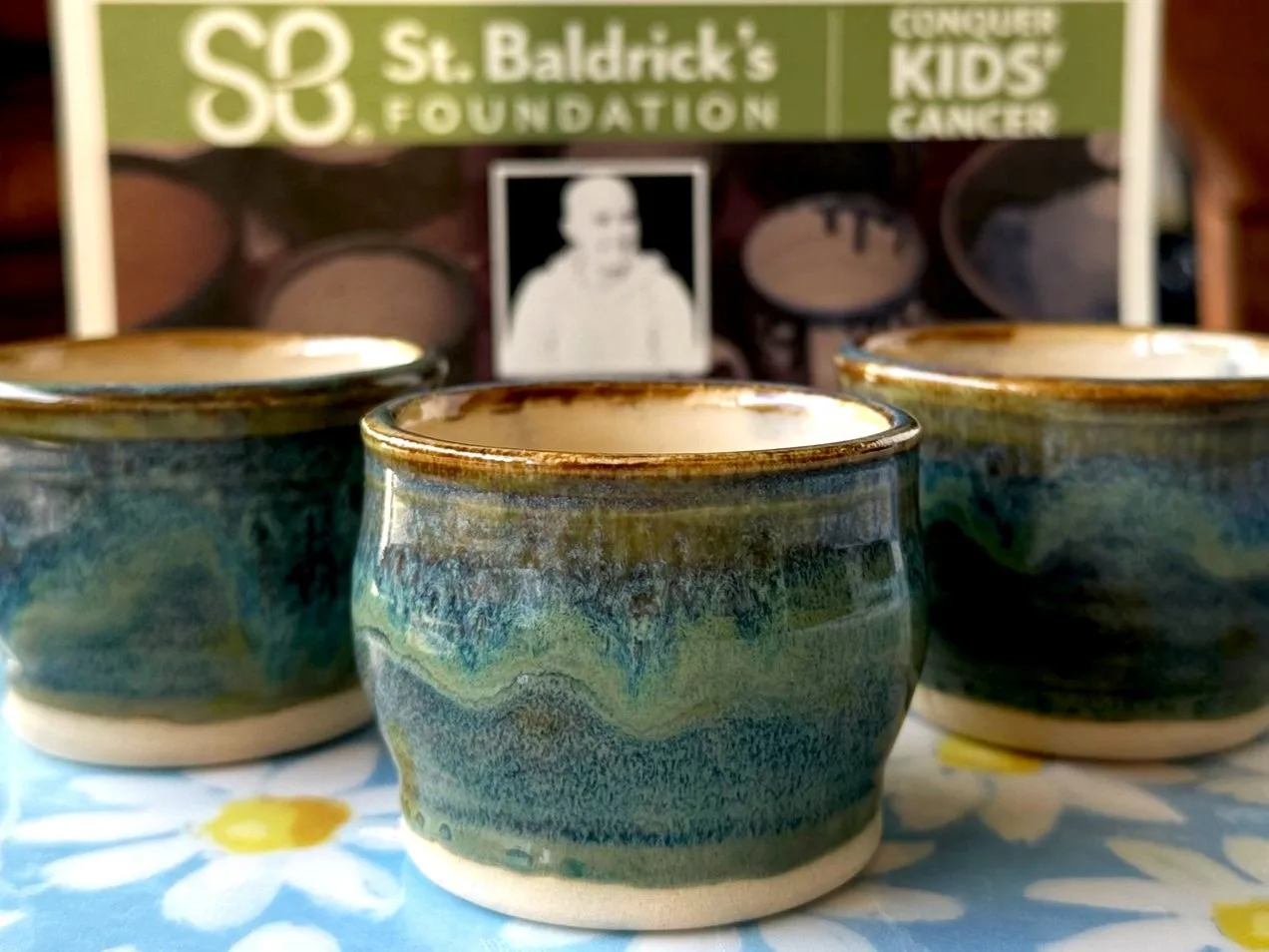 Three small ceramic cups with a variegated blue and green glaze sit on a floral patterned cloth.