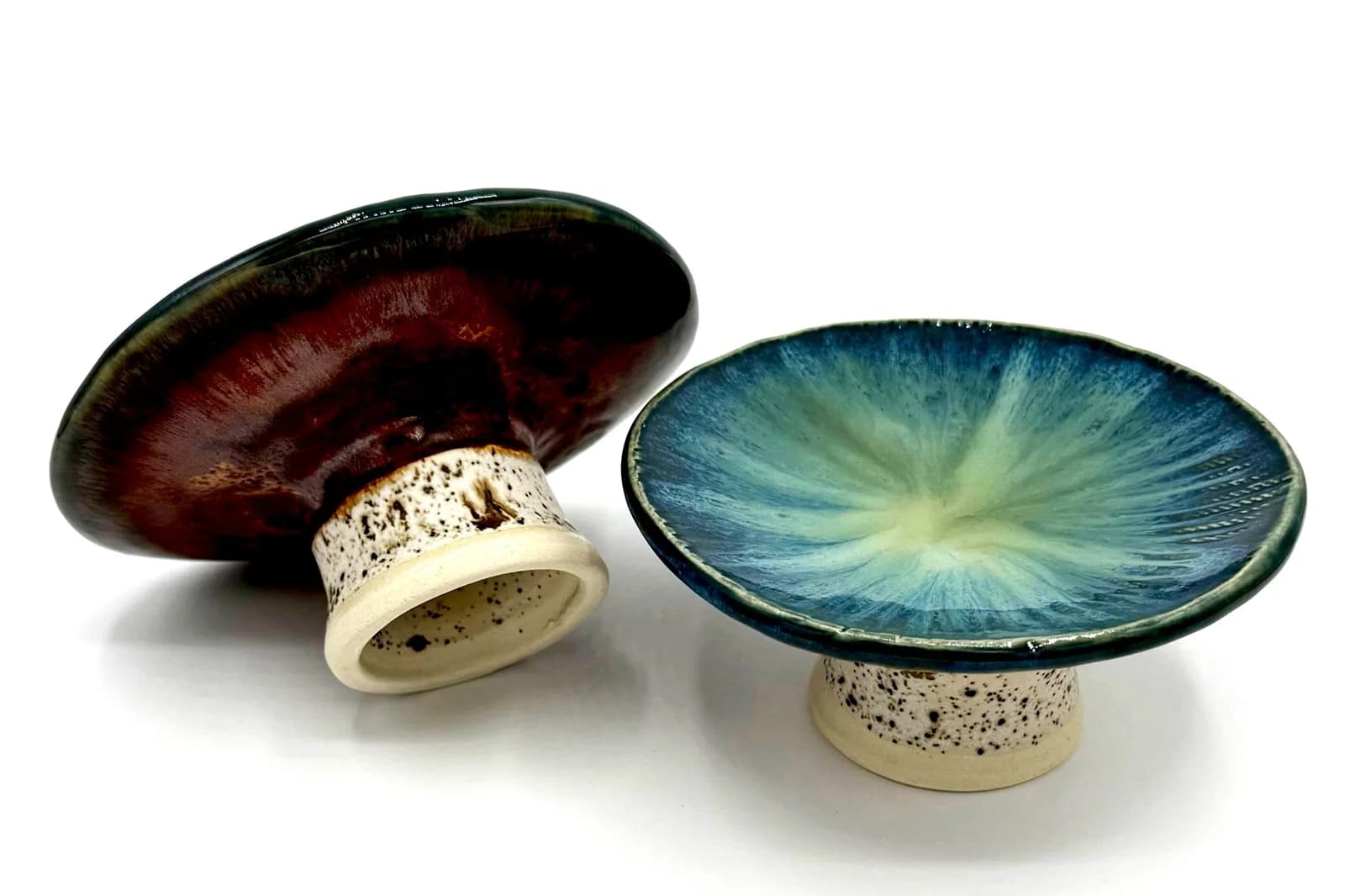 Two ceramic pedestal dishes with glossy glazes in deep red and blue-green hues.