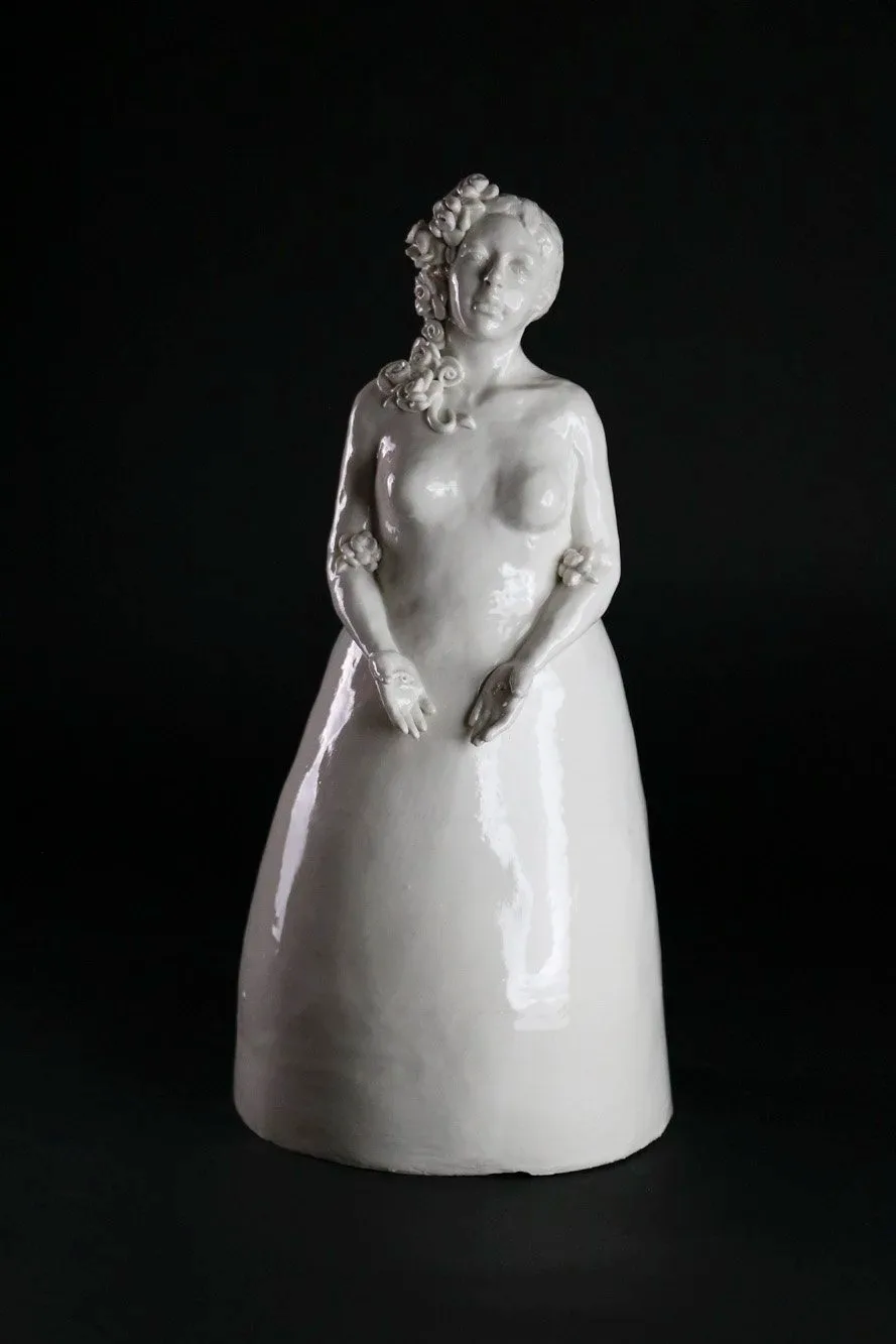 A white ceramic sculpture of a woman in a flowing dress adorned with flowers, presented against a dark background.
