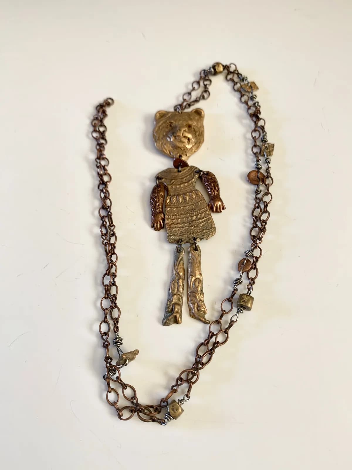 A folk art metal necklace features a bear figure with articulated limbs, crafted from bronze and copper tones.