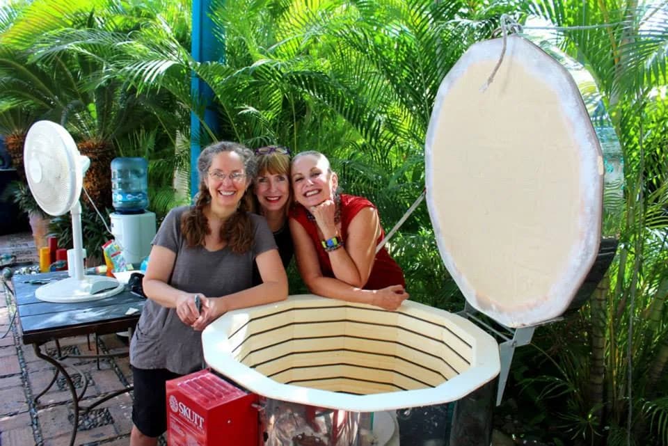 Three artists pose by a large kiln in a lush outdoor studio setting.