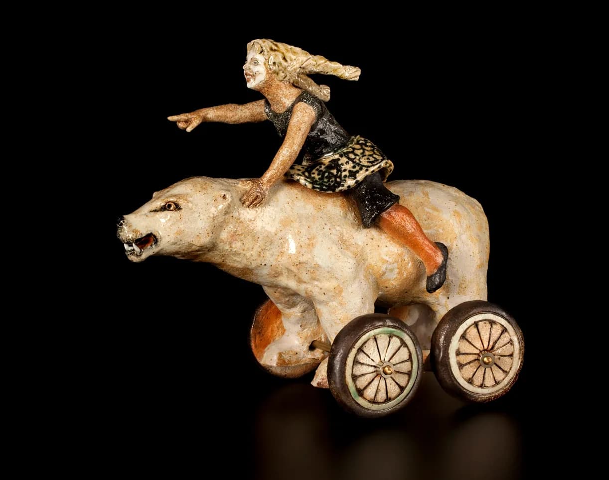 A whimsical ceramic sculpture depicts a girl riding a bear on wheels, rendered in a folk art style.