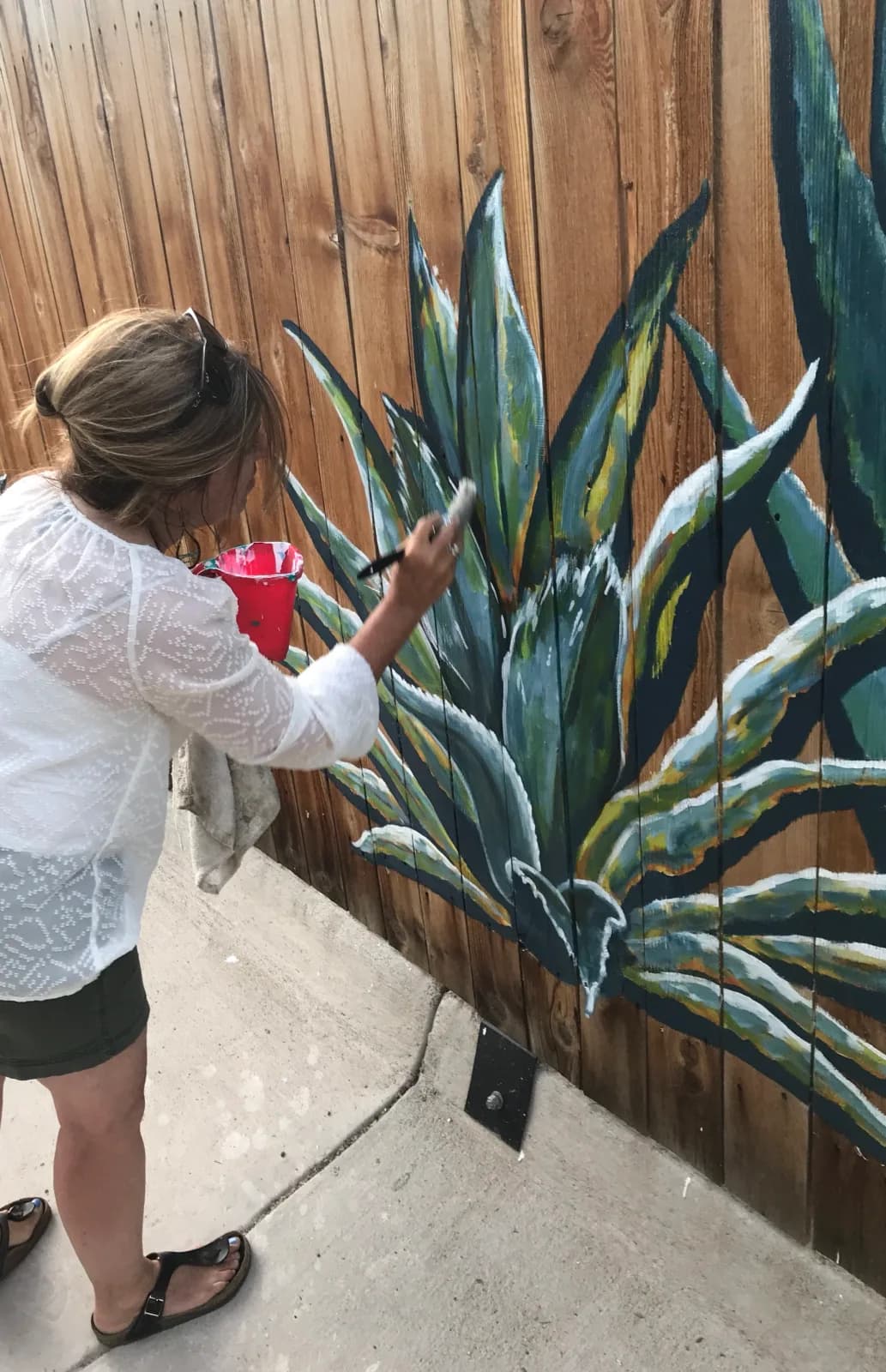 An artist paints an agave mural on a wooden fence, holding a brush and red bucket.