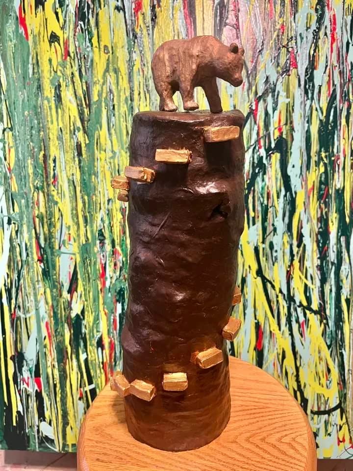 A clay sculpture of a bear atop a log with gold accents, set against a vibrant abstract background.
