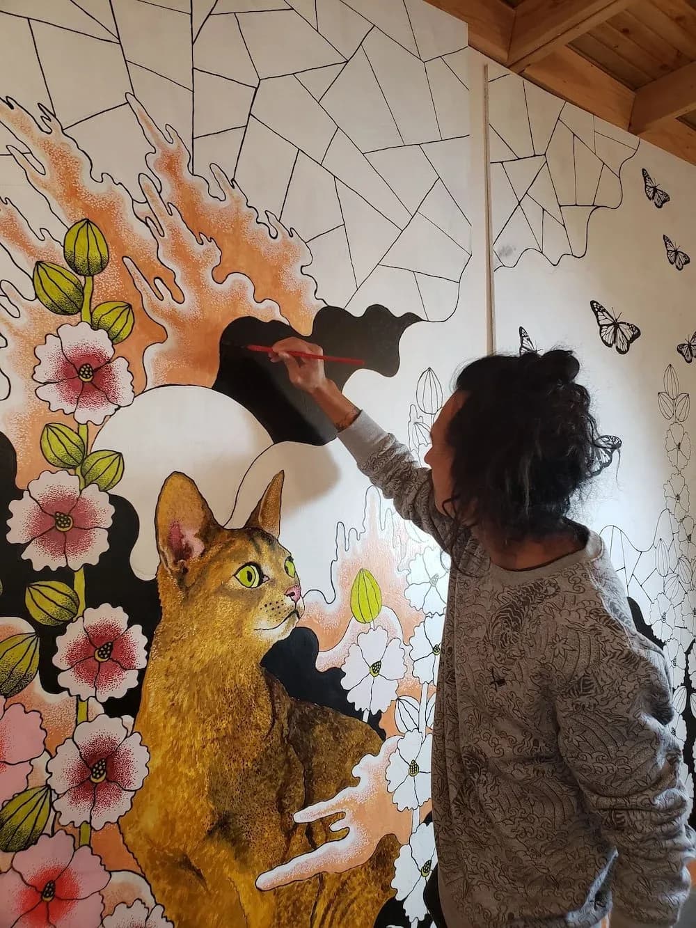 An artist paints a mural featuring a cat and floral elements, using oil and acrylic paints.