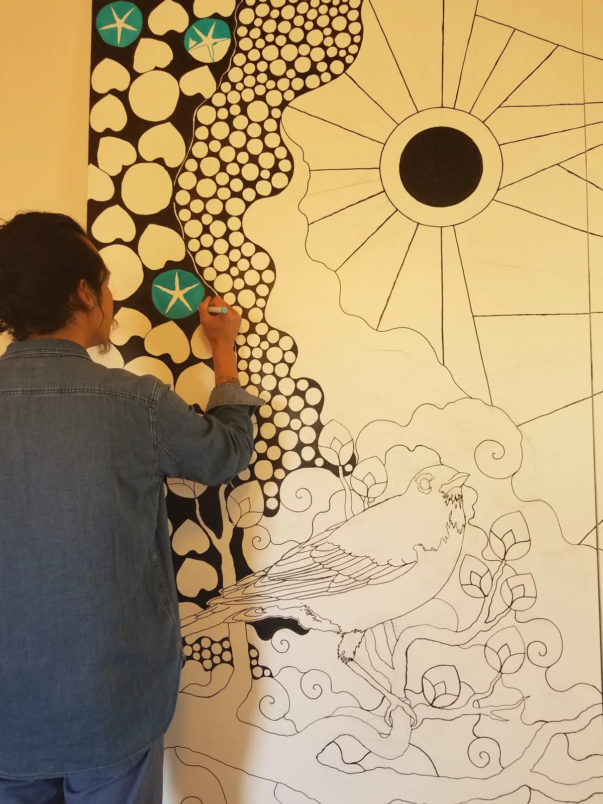 An artist works on a large mural featuring a bird and a sun motif, rendered in ink.