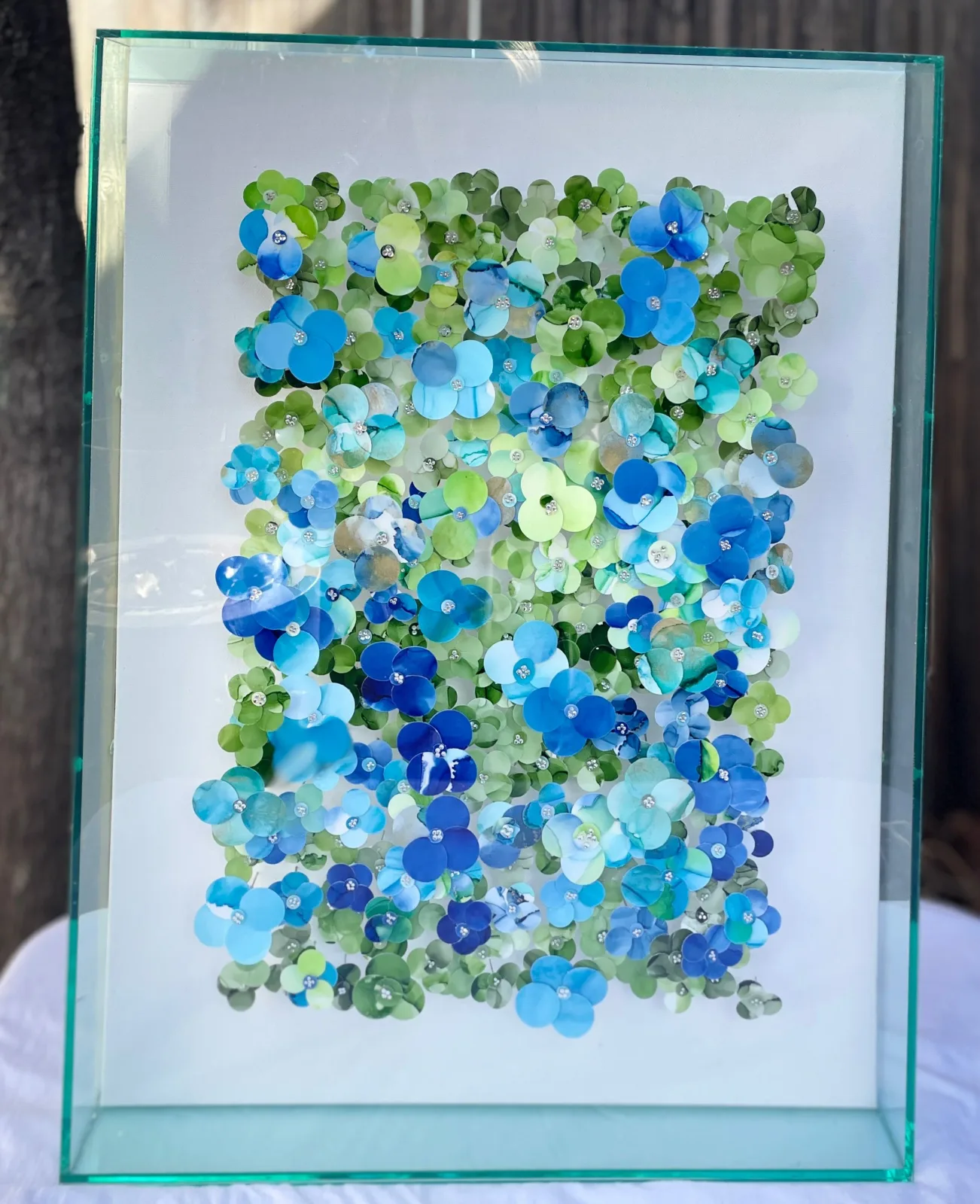 A mixed media artwork featuring numerous blue and green floral elements with embellishments, presented in a glass frame.