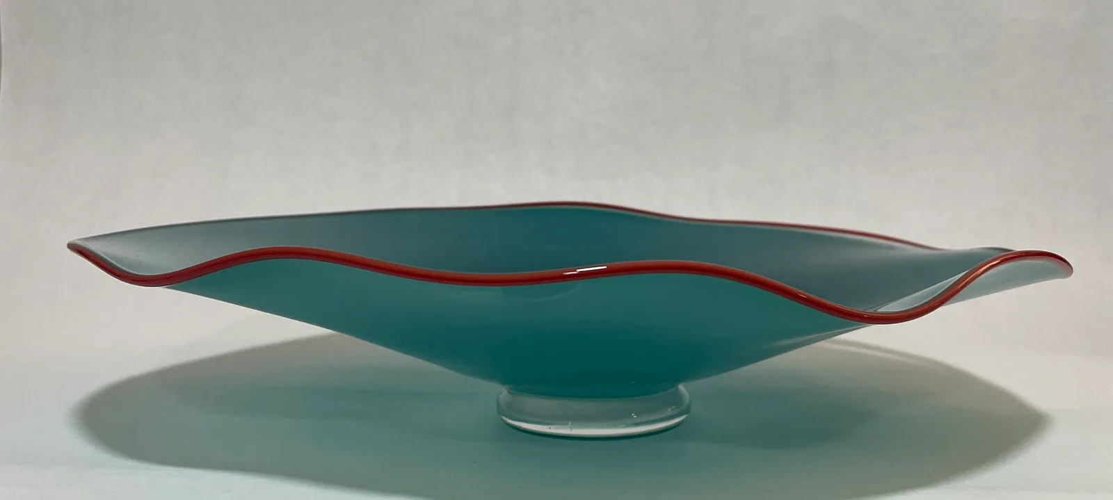 A teal art glass bowl with a red rim rests on a clear glass base.