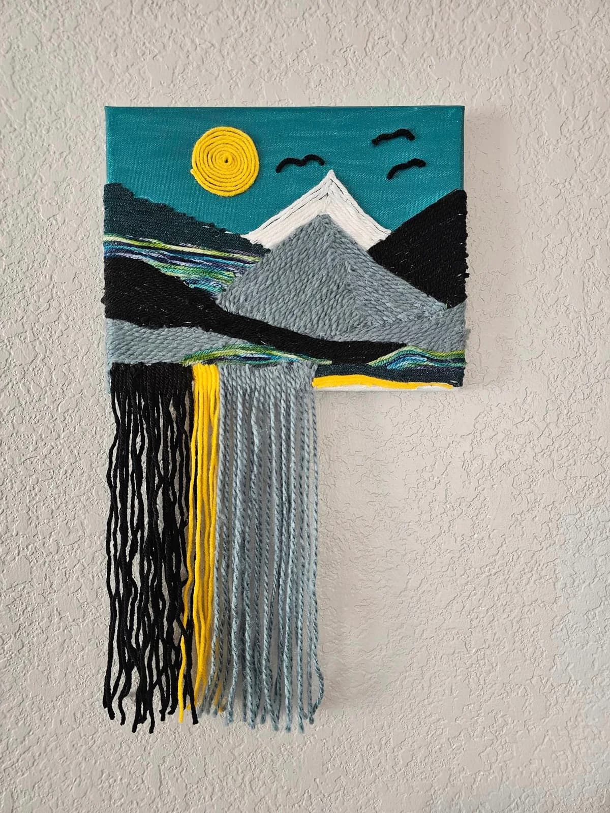A fiber art landscape depicts a sunlit mountain scene with a waterfall, rendered in yarn on canvas.