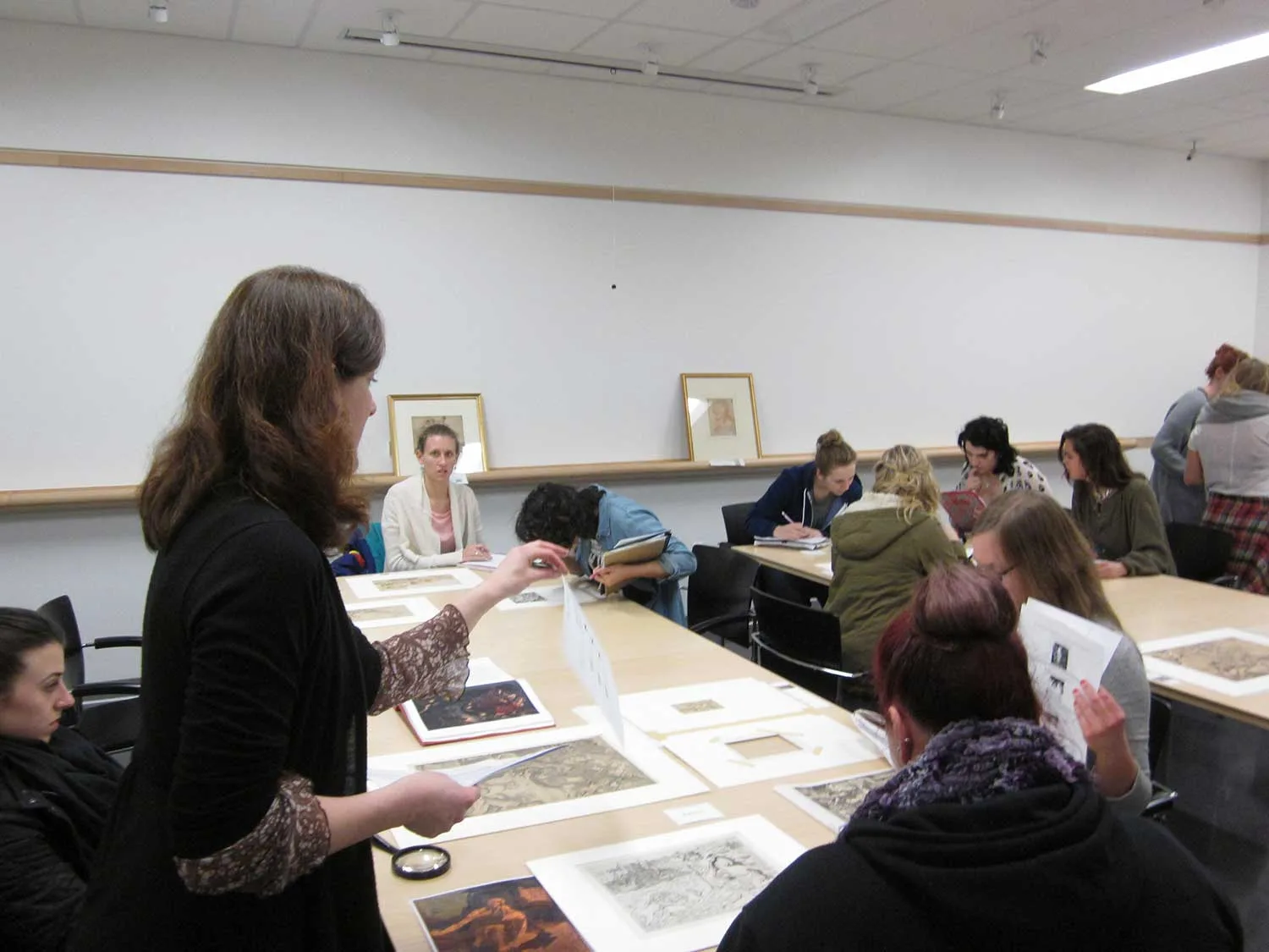 An instructor leads a class in reviewing artworks laid out on tables.