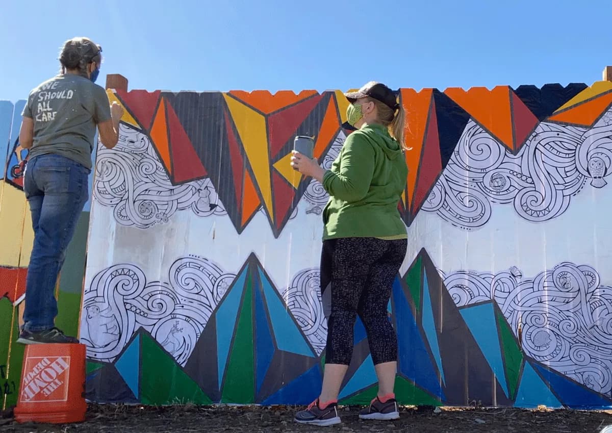 Two artists work on a colorful mural featuring geometric shapes and swirling patterns on a fence.