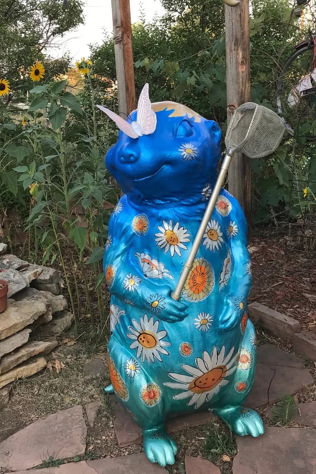 A whimsical blue gopher sculpture adorned with daisies and a butterfly, holding a butterfly net, rendered in a folk art style.