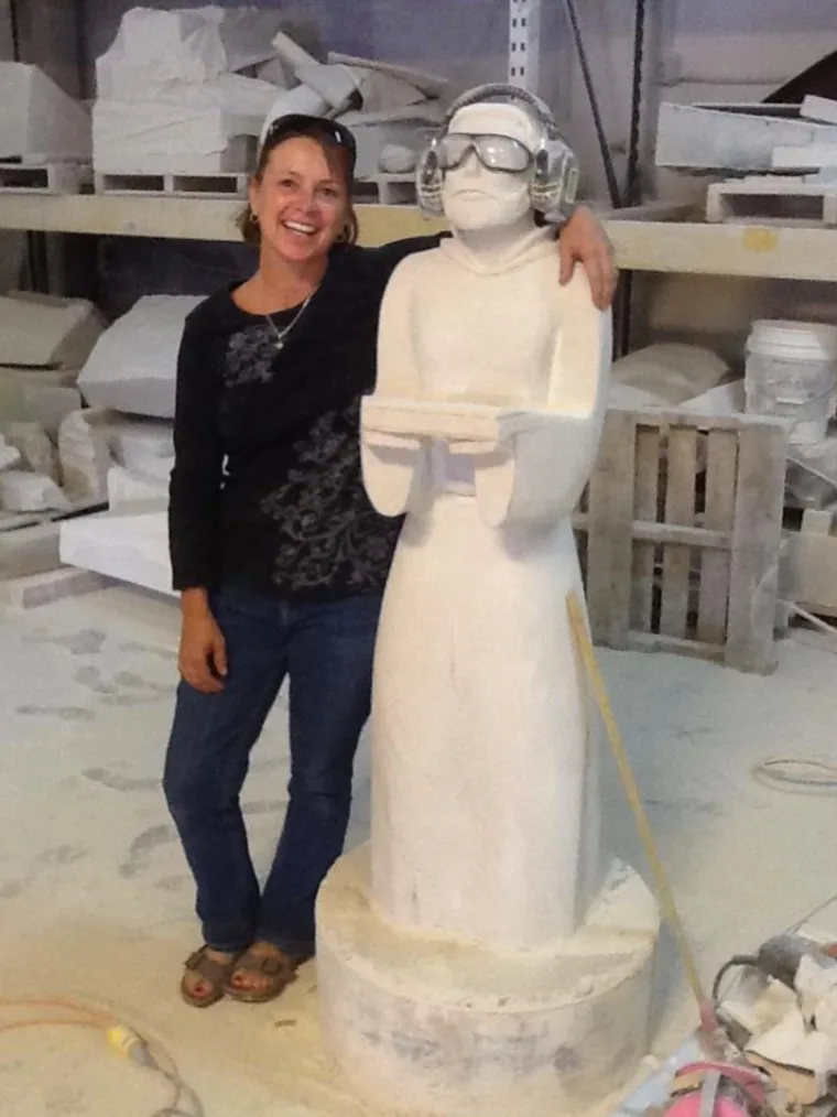 An artist poses with a stone sculpture in progress in a studio setting.