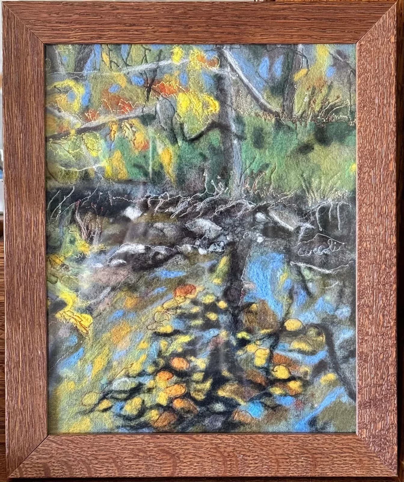 A felted textile artwork depicts an autumn forest scene with a stream and fallen leaves, rendered in vibrant yellows, blues, and greens.