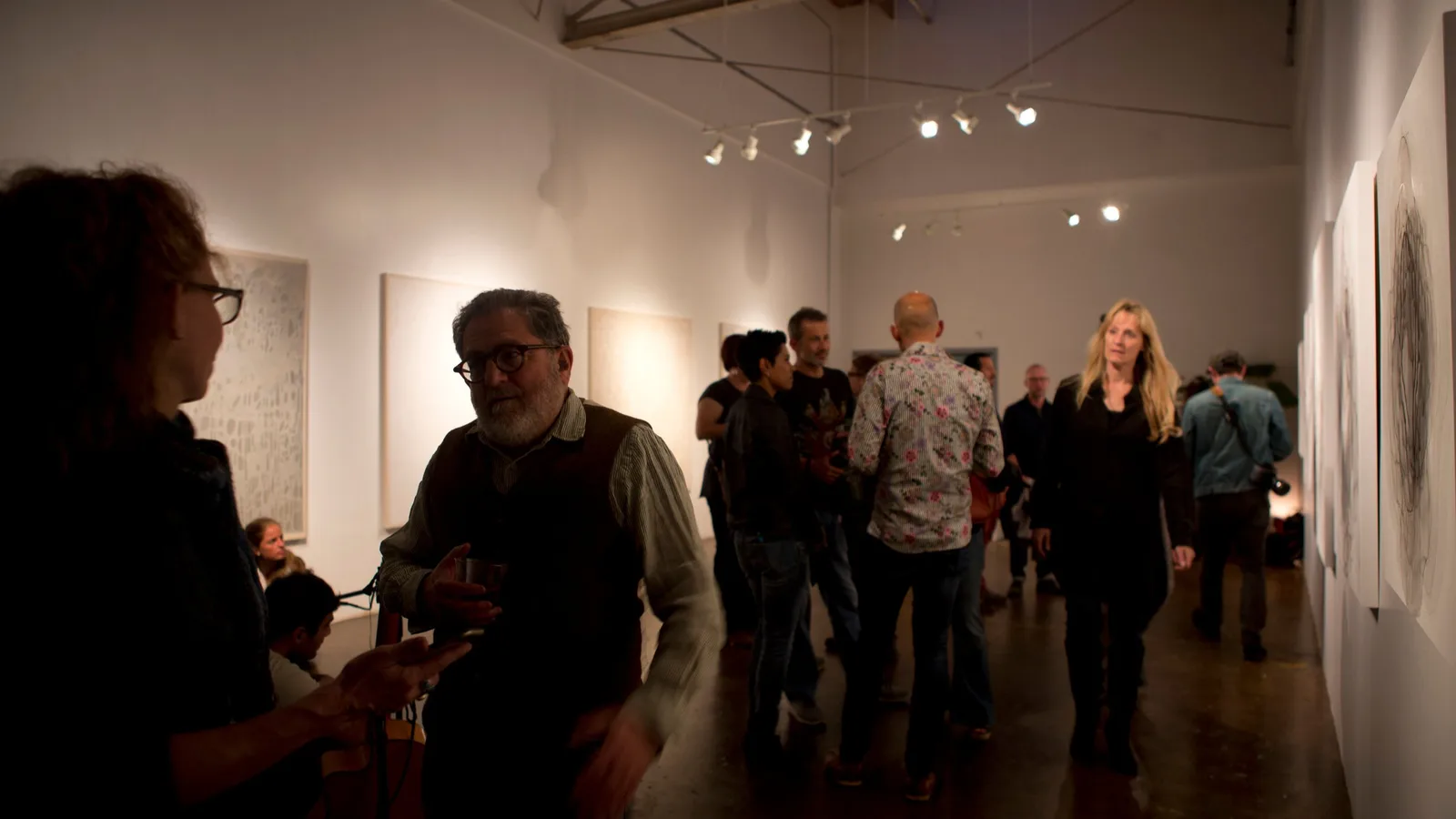 People mingle at an art exhibition, with artwork displayed on the walls.