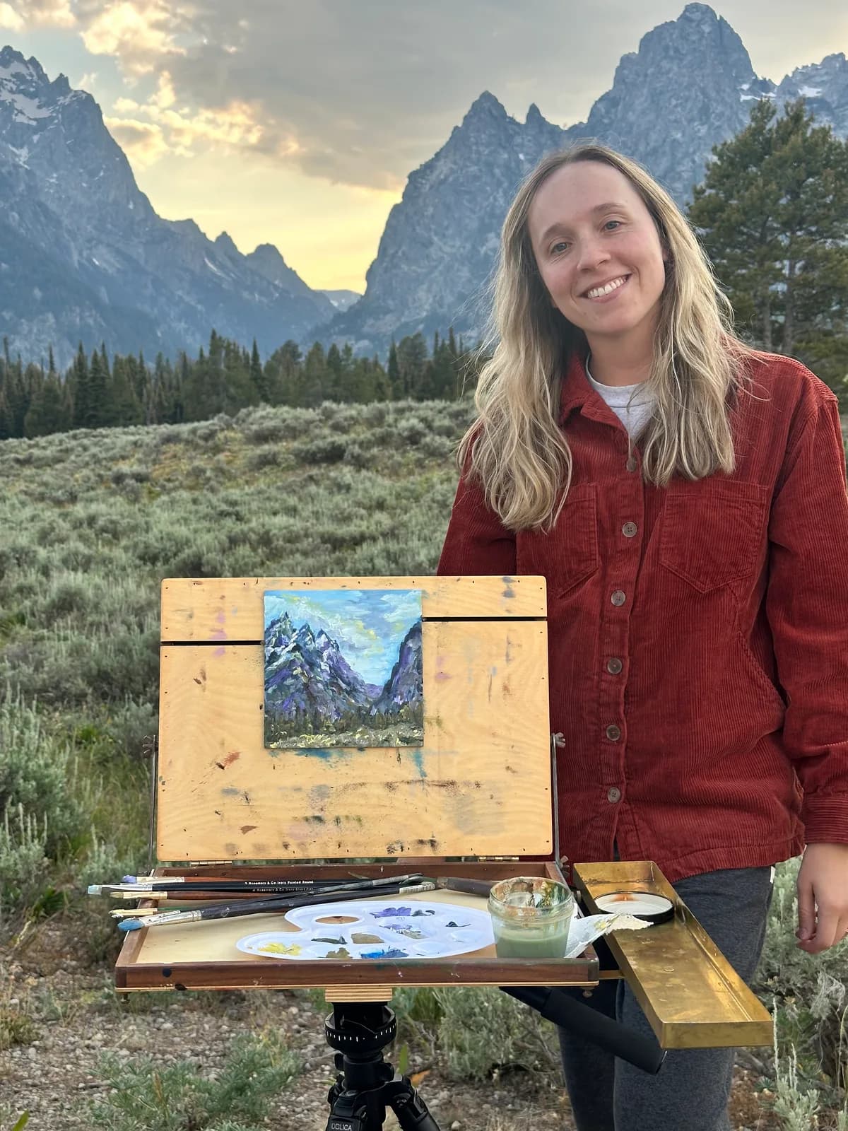 An artist paints a mountain landscape en plein air with oil on an easel, featuring a palette with Heath and Tide colors.