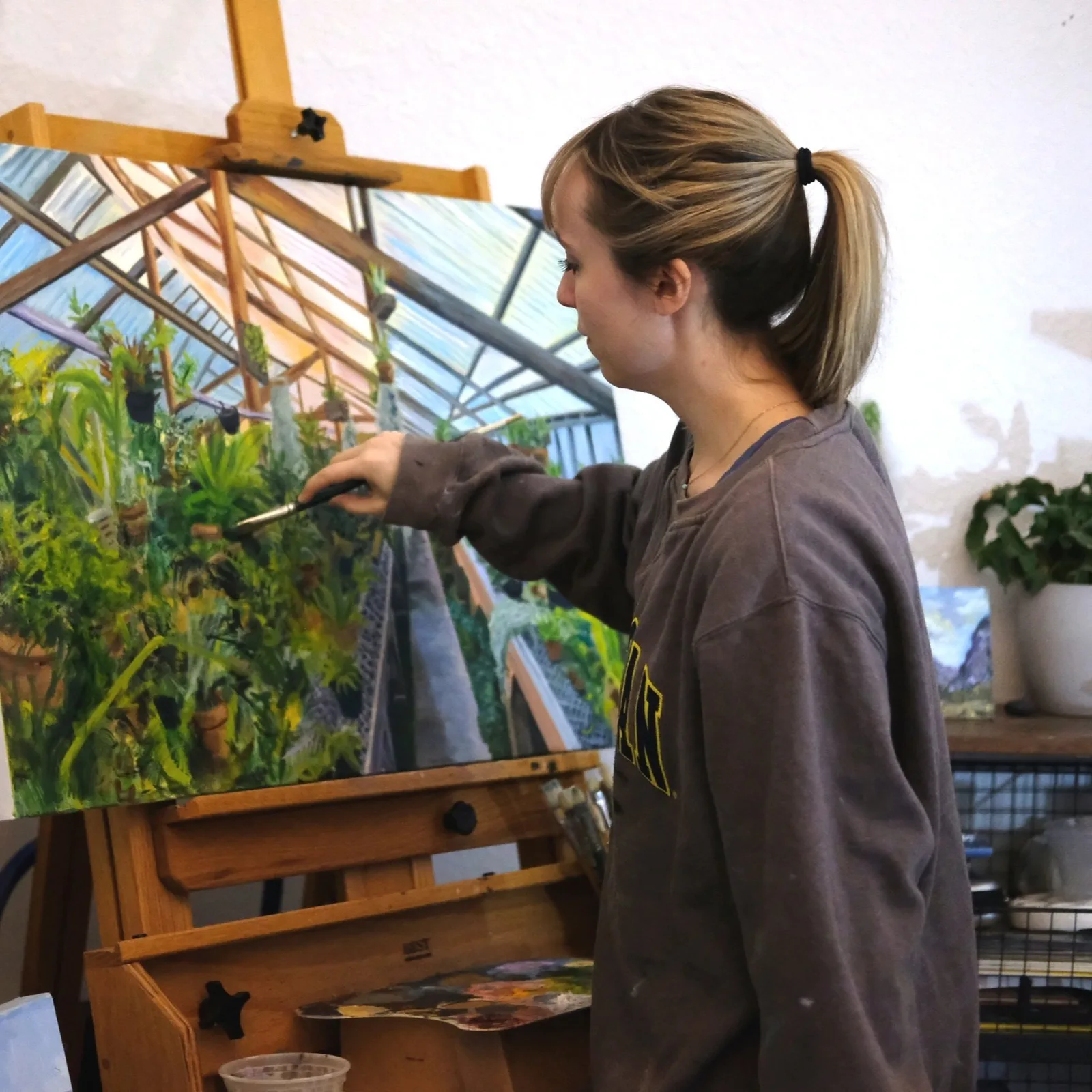 An artist paints a lush greenhouse scene on canvas in their studio.