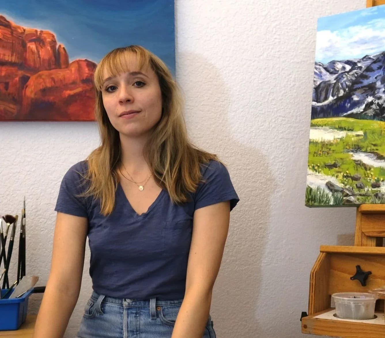 An artist in her studio poses with two landscape paintings, one of red rock formations and another of mountains.