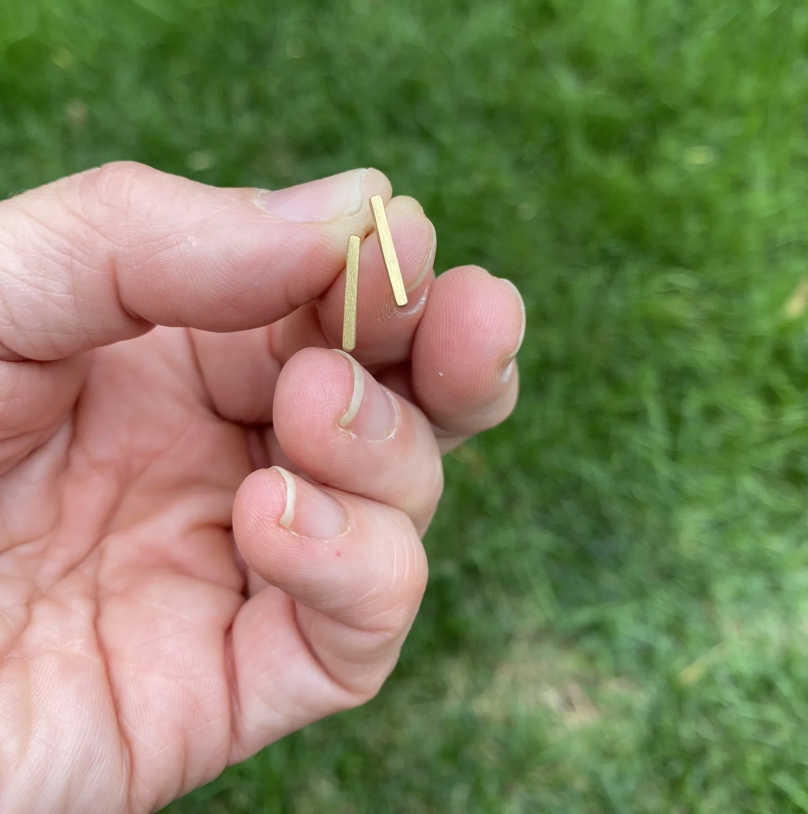 Minimalist gold bar earrings presented by hand against a grassy background.
