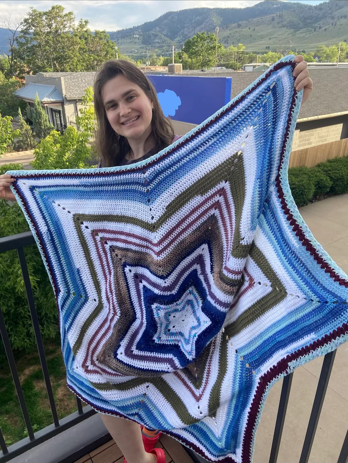 A woman holds a star-shaped crochet blanket with stripes of blue, white, and earth tones, displayed on a balcony with a mountain view.