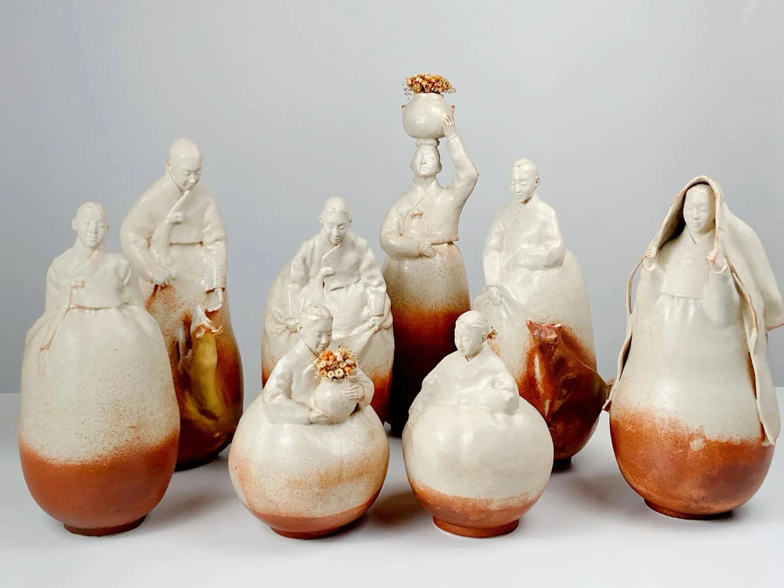 A collection of ceramic figures in traditional Korean clothing, holding flowers and pots, with a nutmeg and gray nickel glaze.