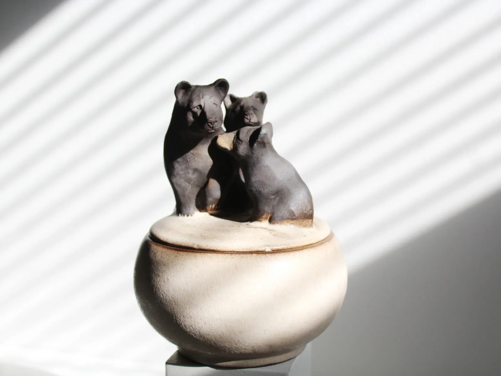 A ceramic sculpture of three bears sits atop a lidded pot, rendered in shades of Wild Sand and Dove Gray.