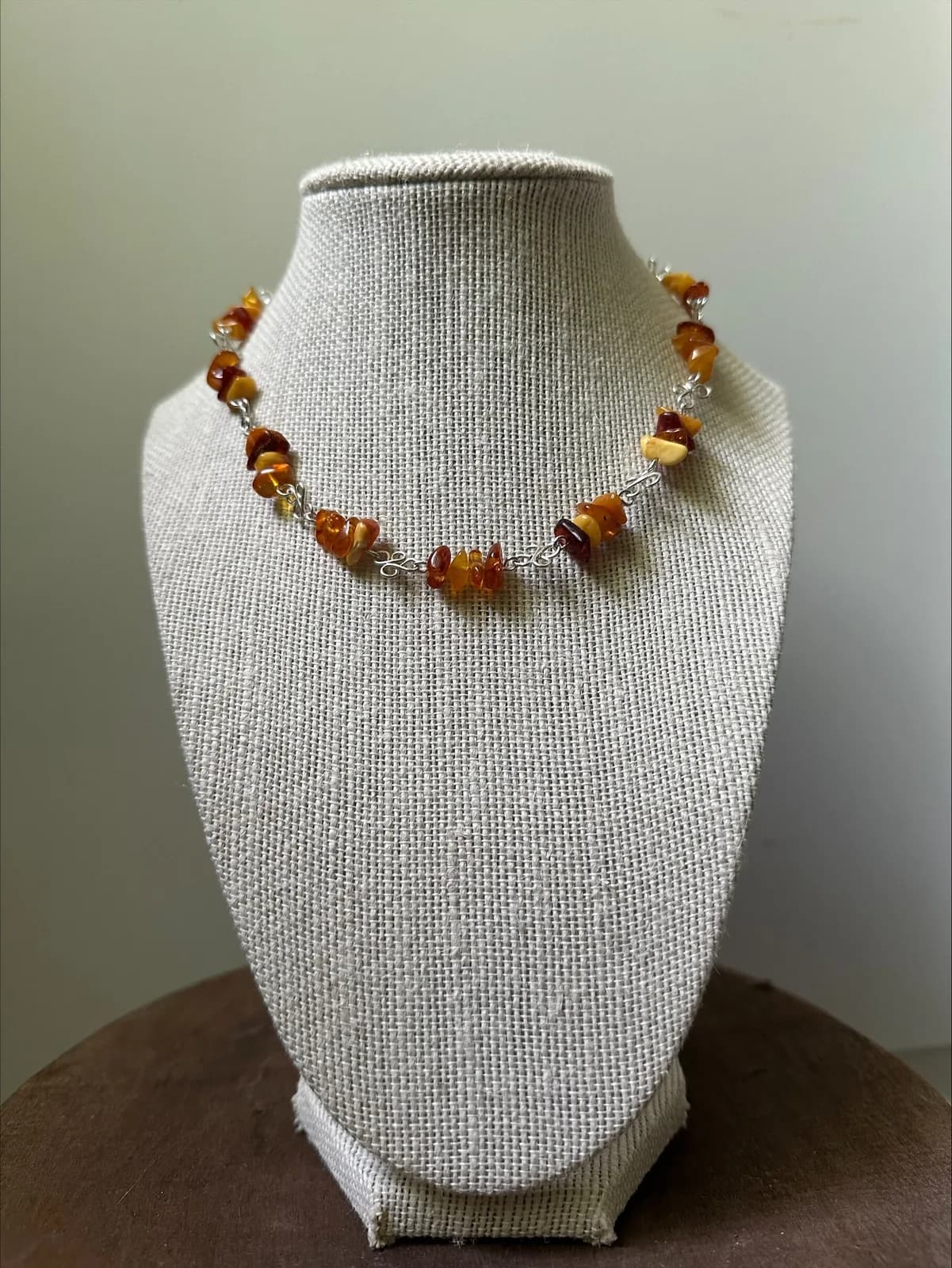 A necklace of amber beads on a silver chain is displayed on a burlap-covered bust.