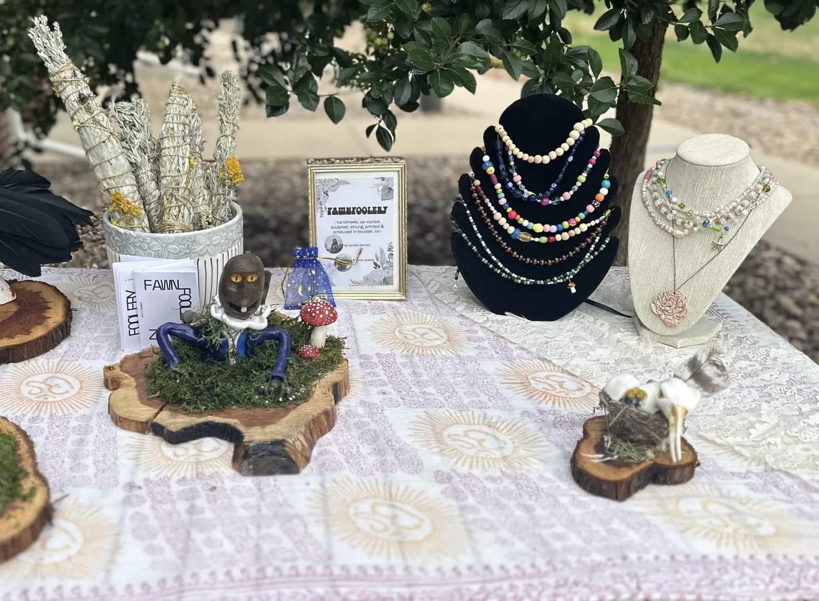 Handmade jewelry and figurines are displayed on a table covered with a patterned tablecloth, set outdoors under leafy branches.