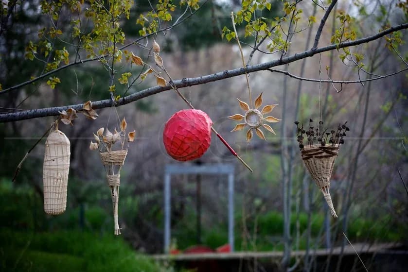 A collection of nature-inspired fiber art ornaments hang from a tree branch, featuring a red sphere and woven baskets.