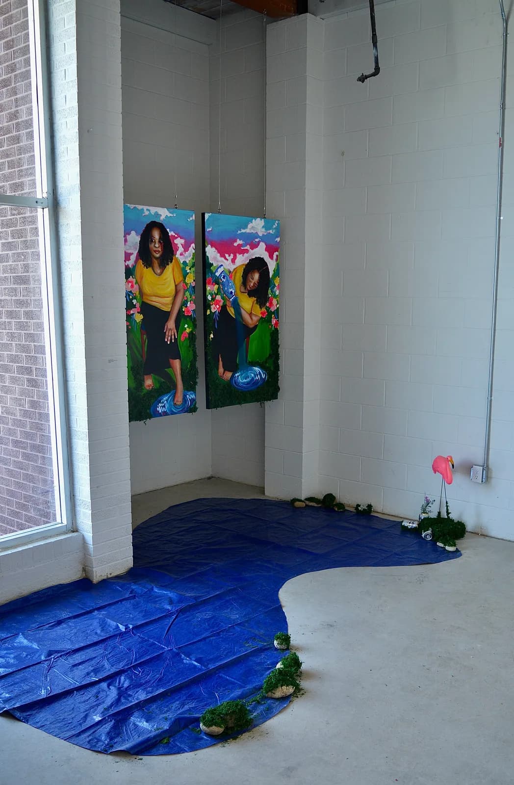 Two paintings of a woman in a yellow shirt with flowers are displayed in a room with a blue tarp representing water and a pink flamingo.