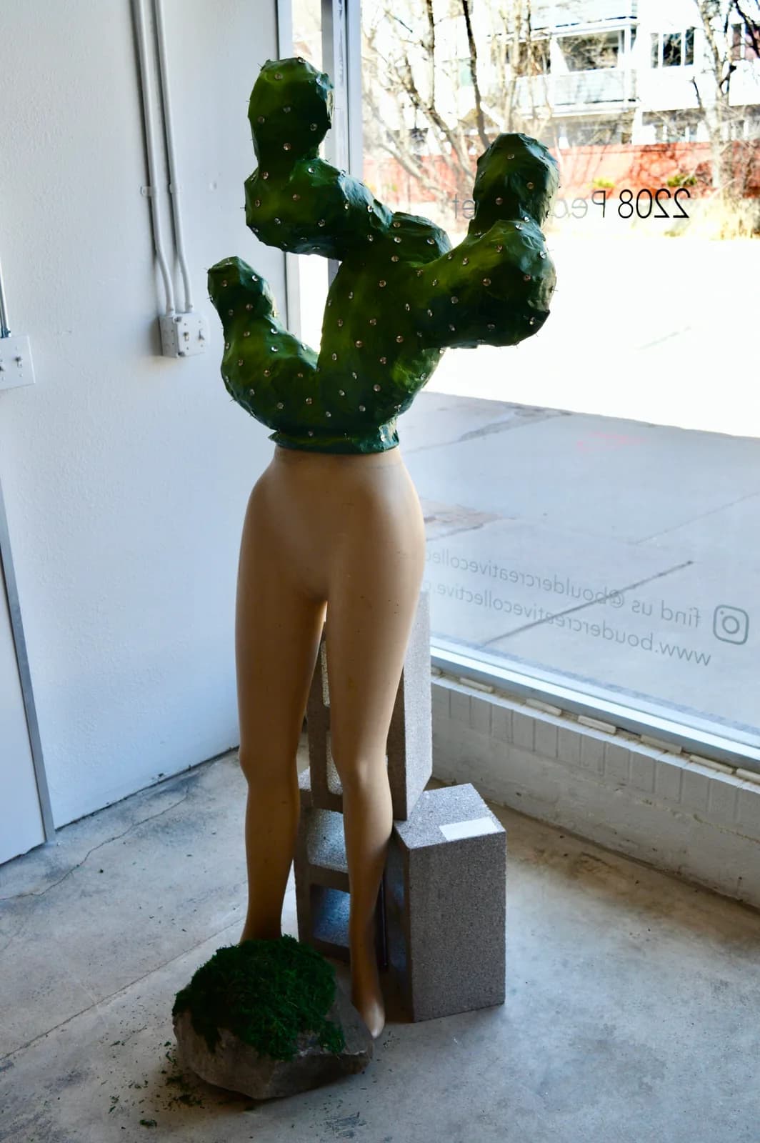 A sculpture of a cactus torso atop mannequin legs, resting on cinder blocks and moss-covered stone, displayed in a storefront.