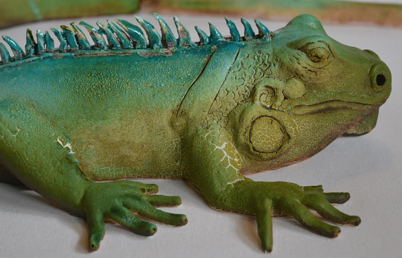 A ceramic sculpture of a green iguana with textured skin and turquoise spines, presented on a neutral surface.
