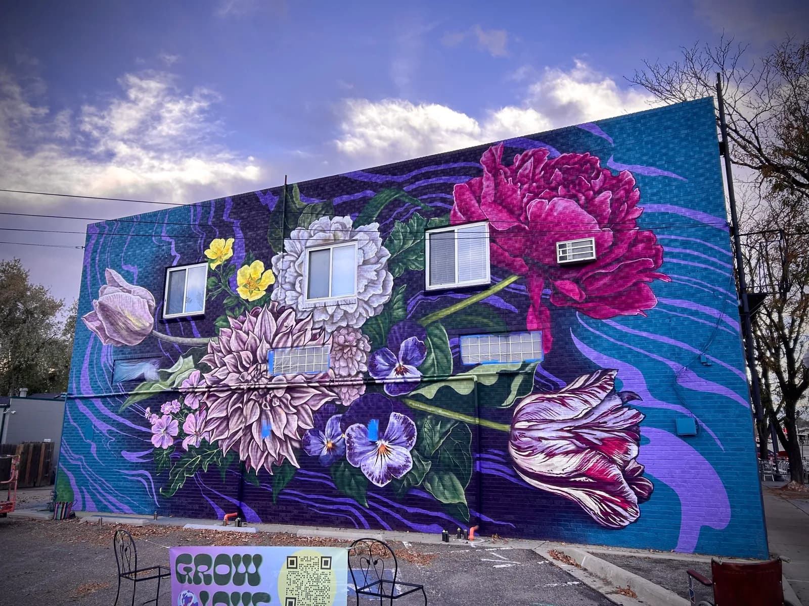 A vibrant floral mural in shades of plum and blue haze adorns a brick building, featuring large peonies and tulips.