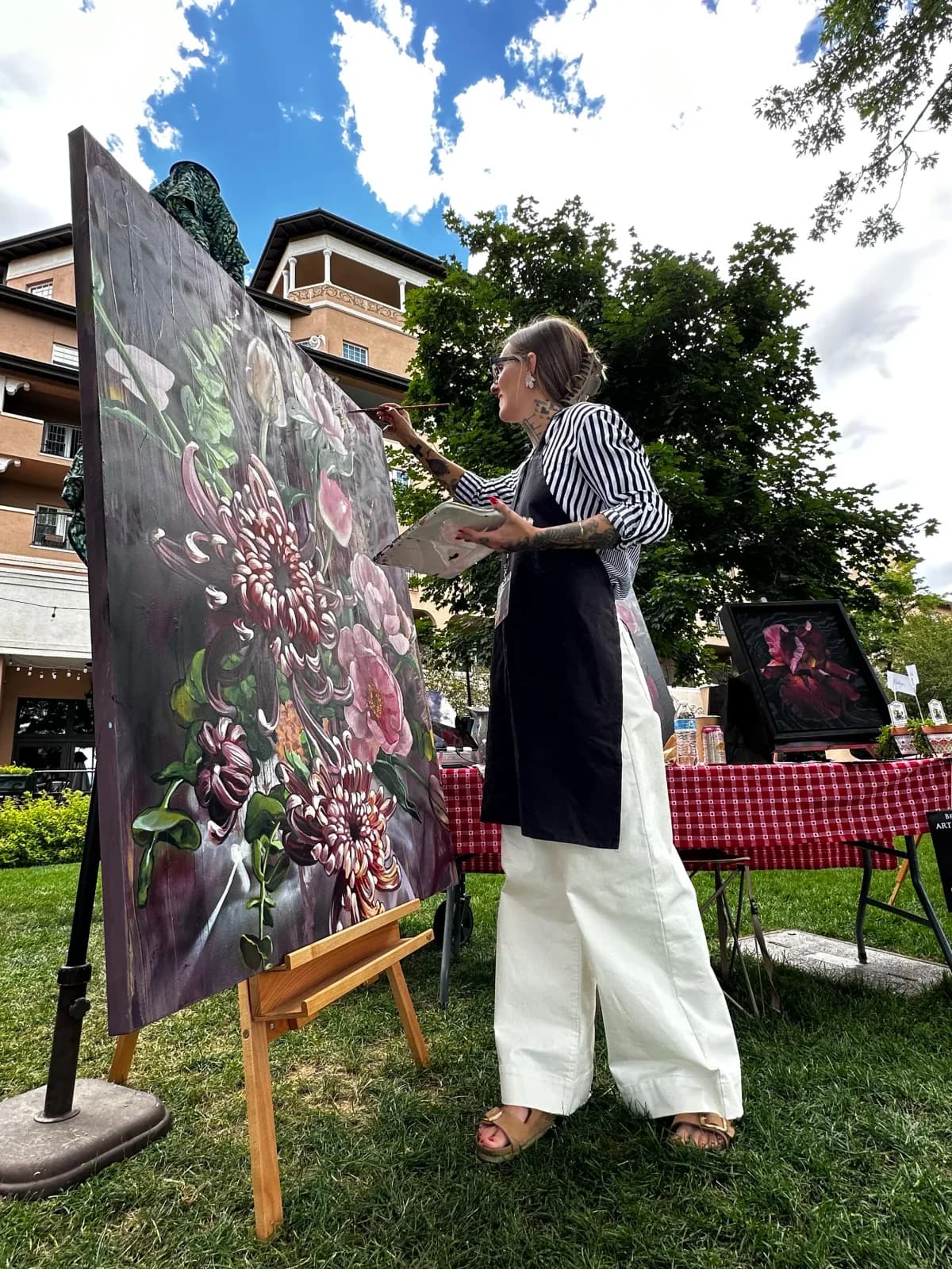 An artist paints a large floral artwork outdoors, working on a canvas with a palette in hand.