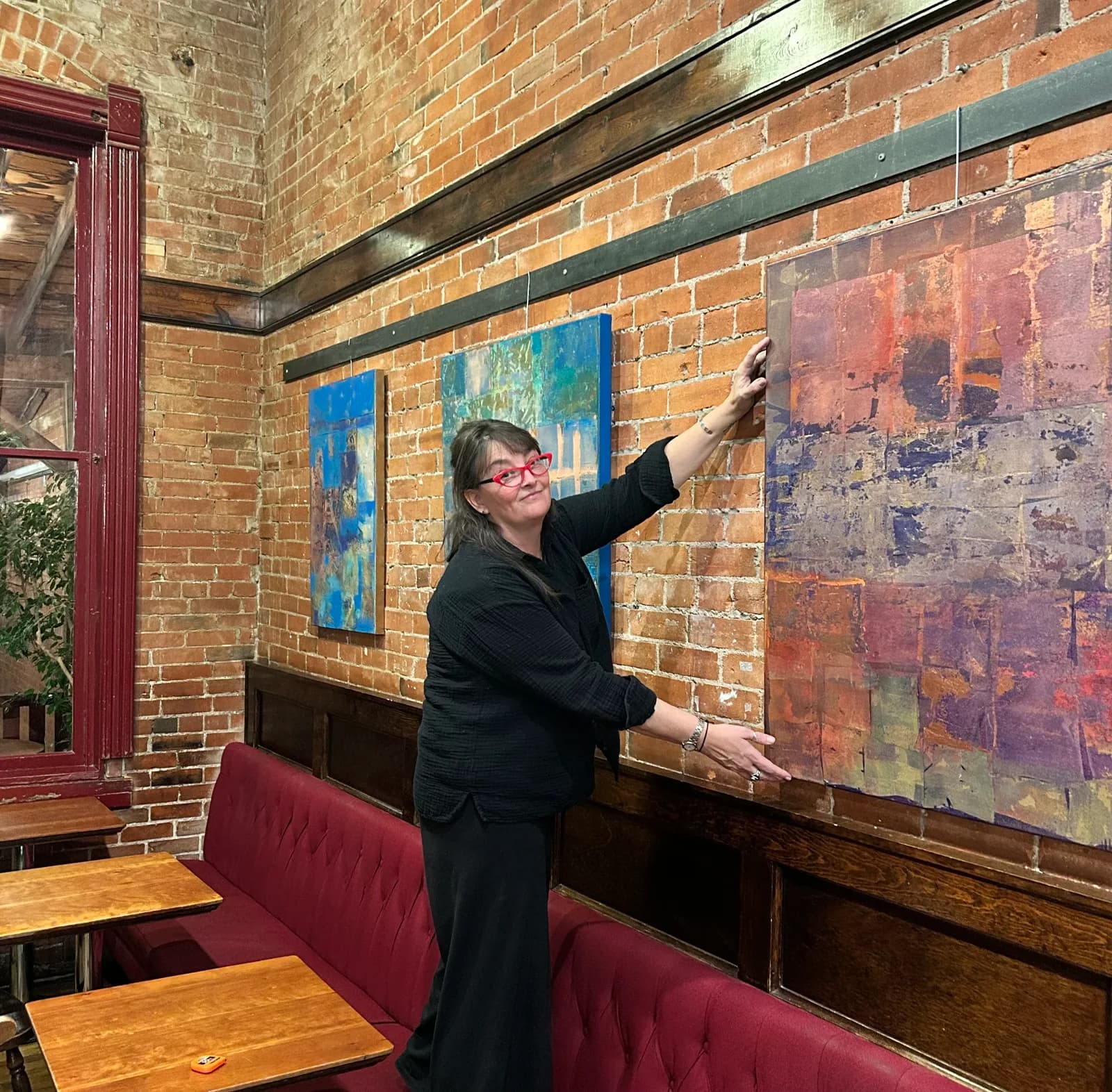 An artist adjusts an abstract painting in a gallery setting with brick walls and red seating.