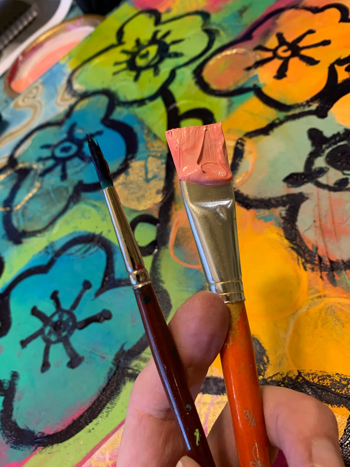 A close-up of two paintbrushes held over a vibrant, abstract floral painting in progress.