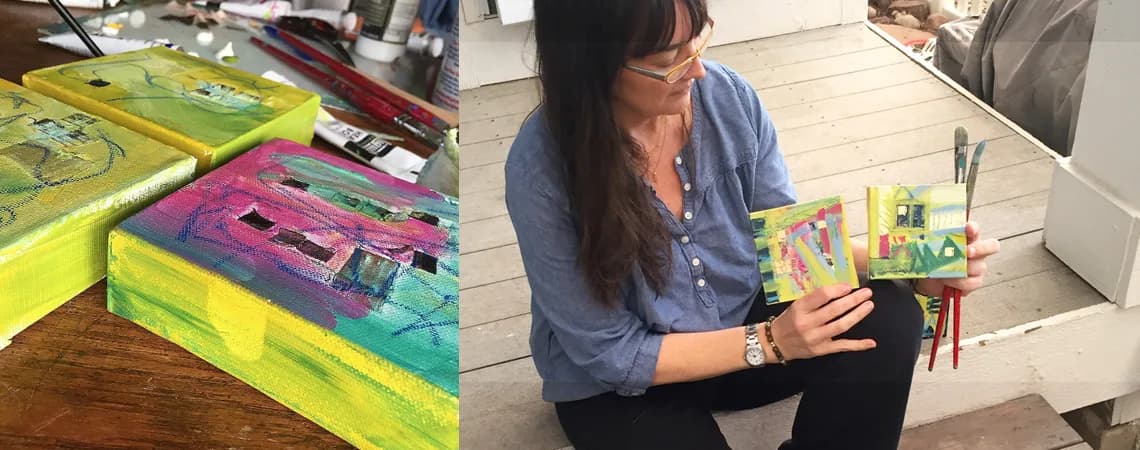 Artist's hands hold small abstract paintings in a studio setting with art supplies.