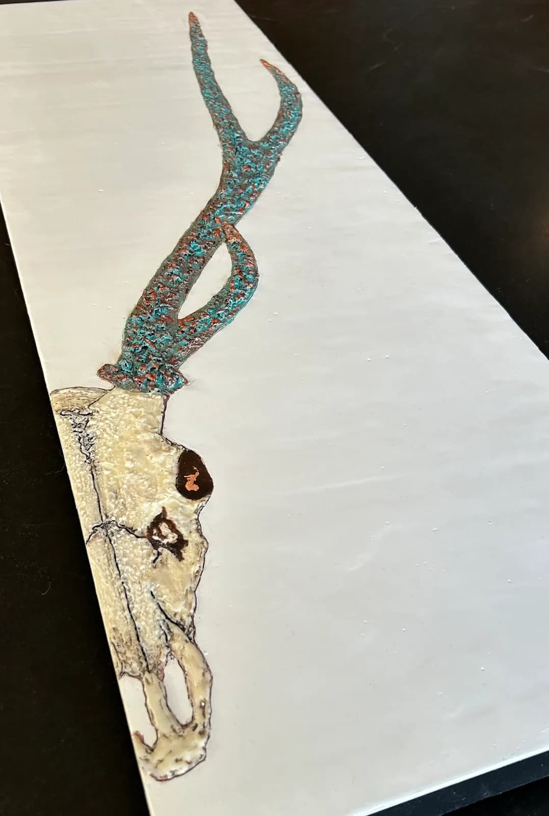 A mixed media artwork depicts a deer skull with textured teal and copper antlers against a white background.