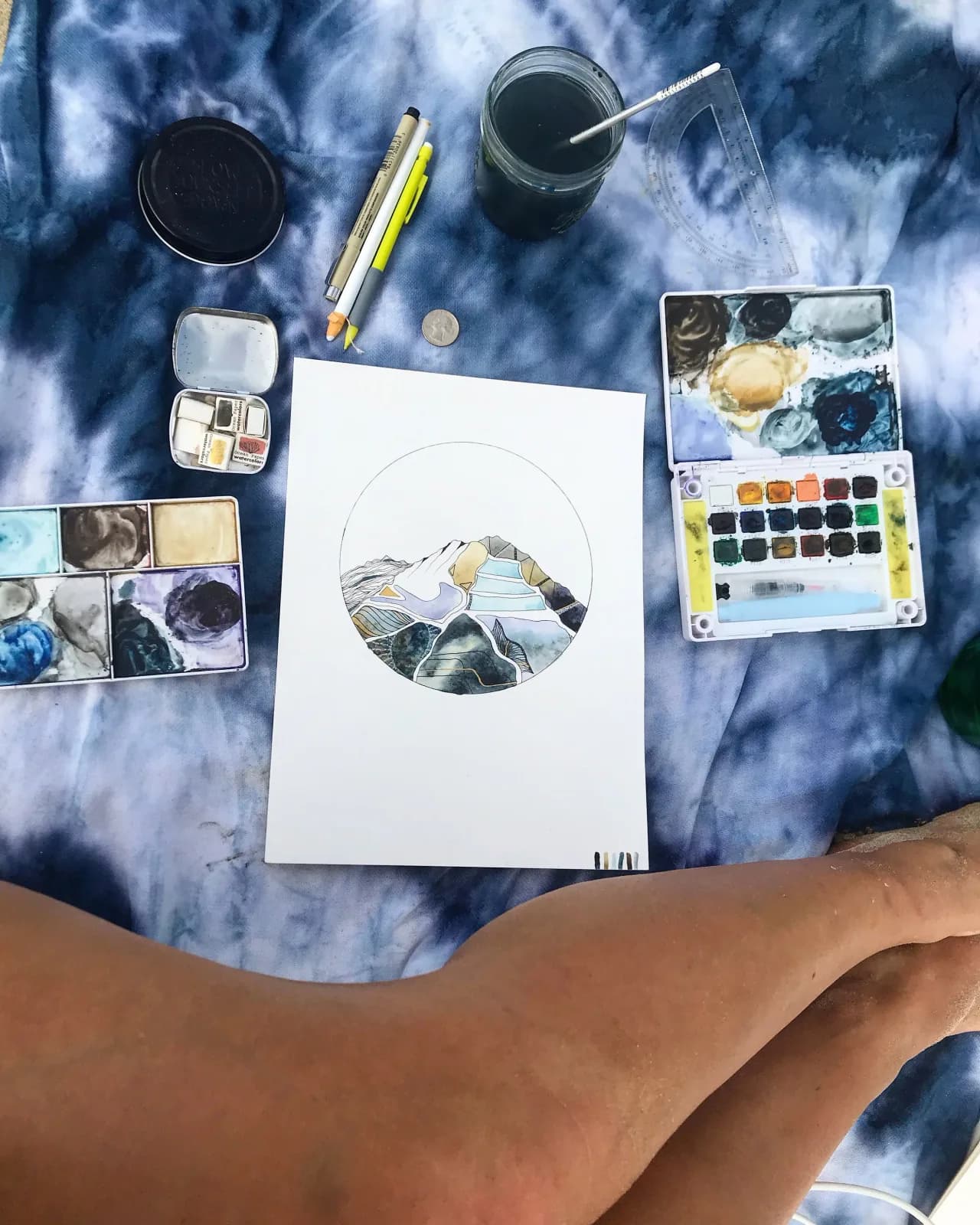 A watercolor painting of mountains in progress, surrounded by art supplies on a tie-dye cloth.