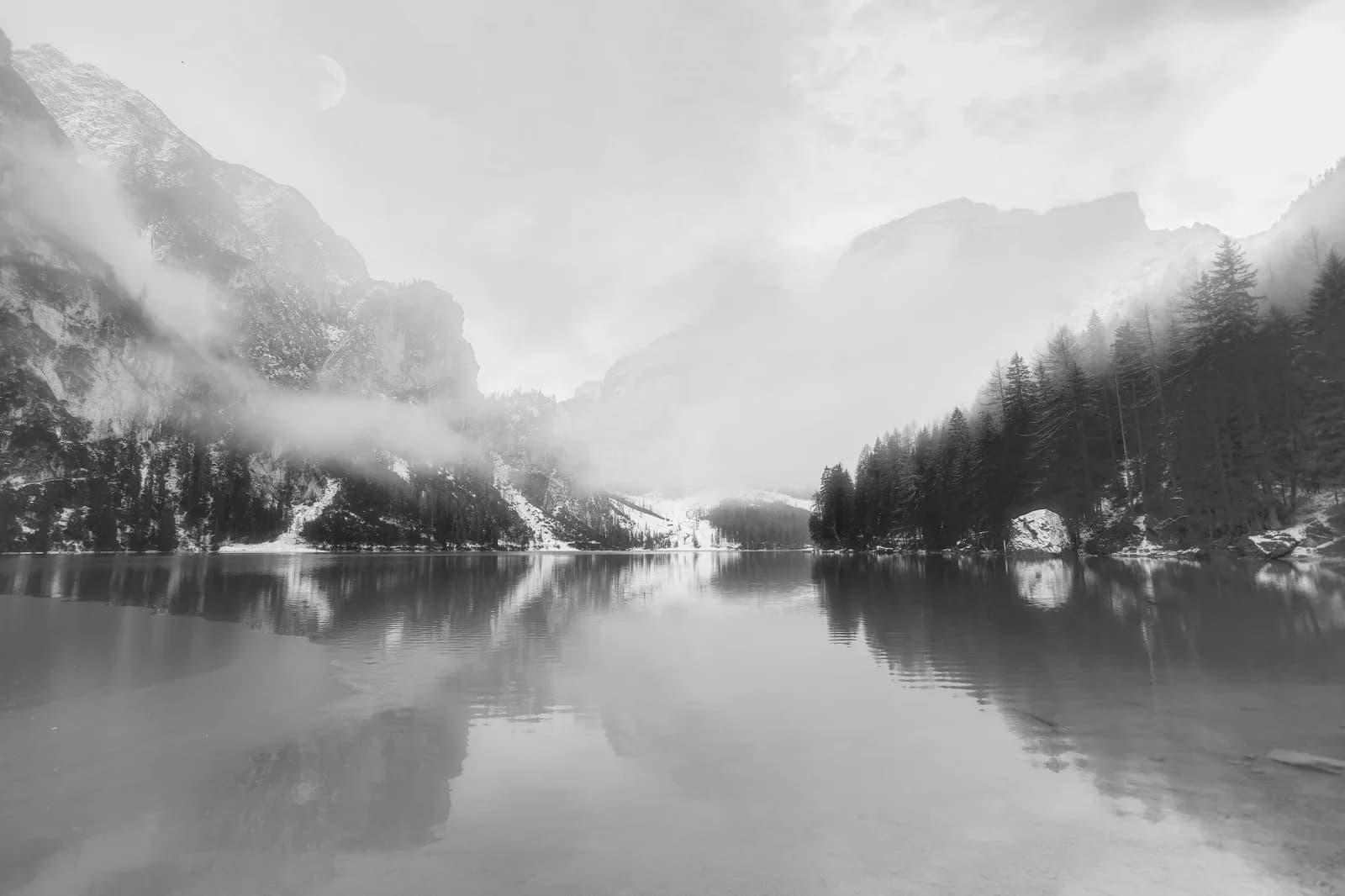 A black and white impressionistic photograph captures a misty mountain lake with reflections of trees and fog-laden peaks.