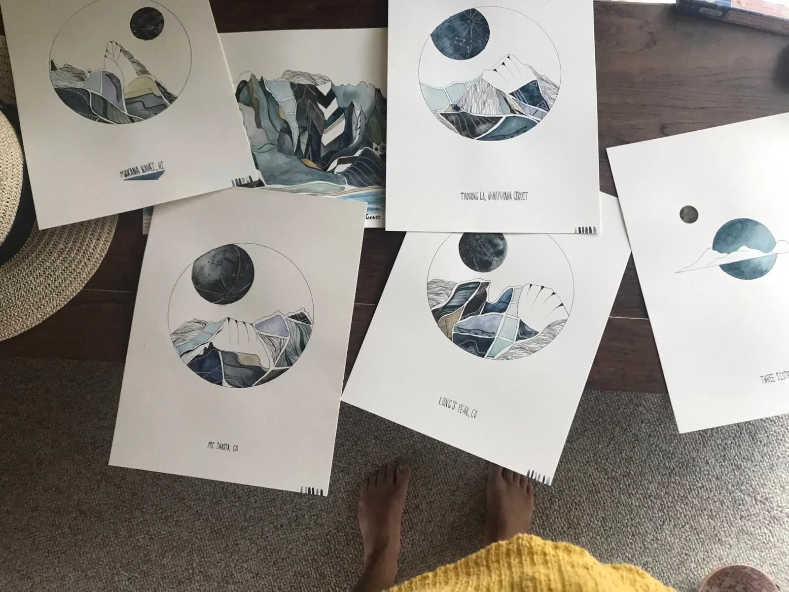 Several watercolor paintings of mountains and moons are laid out on a table.